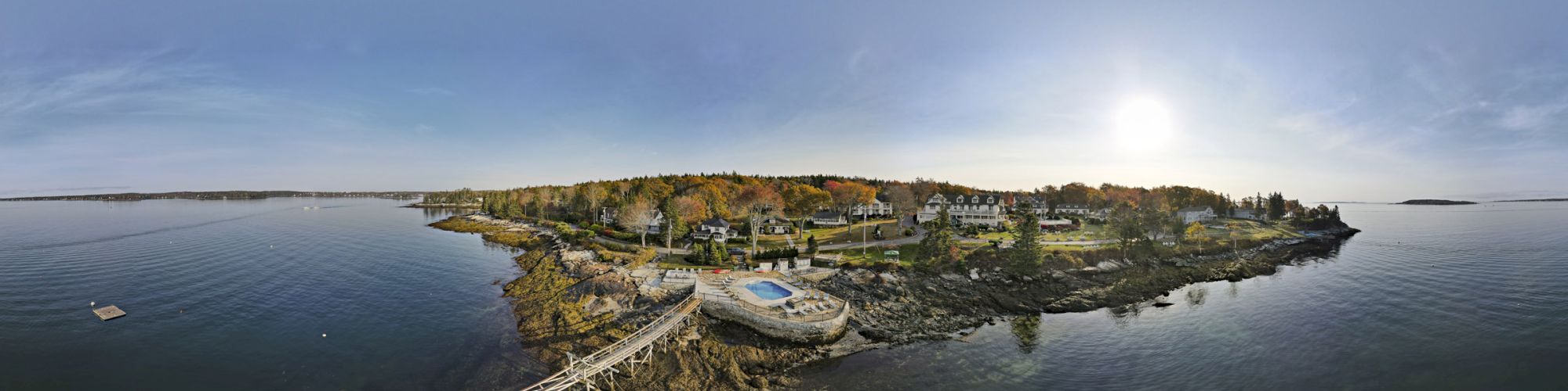 Aerial view of a scenic coastal area with a pier, rocky shore, houses, and lush trees under a bright sky, and the sun low in the sky, ending the sentence.