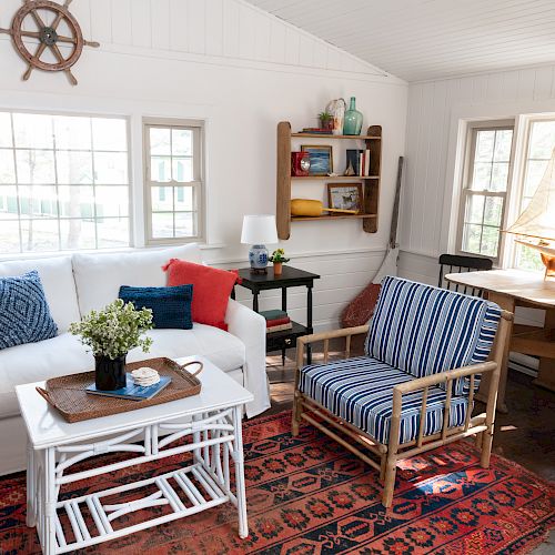 A cozy living room with a nautical theme features a white couch, striped chairs, a rug, a ship model, and a table by the window with chairs.