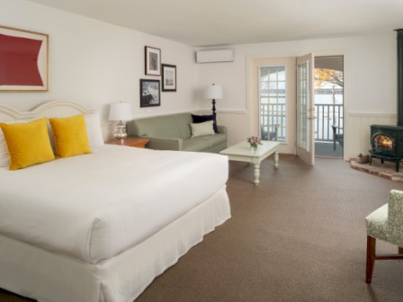 A spacious bedroom with a large bed, yellow cushions, a green sofa, coffee table, and a wood-burning stove. Outdoor view through open doors.