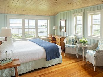 A cozy bedroom with wooden floors, a bed, two chairs, a desk, and a view of the water through three large windows, ending the sentence.
