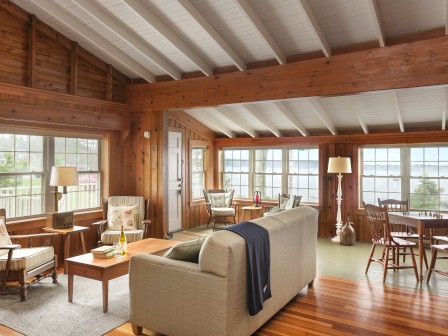 A cozy, wooden living room features a beige sofa, armchairs, coffee table, and dining area with large windows displaying a scenic view.