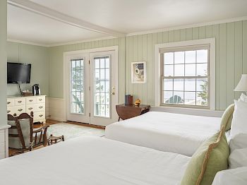 The image shows a cozy bedroom with two beds, a dresser, a TV, a spacious window, and French doors. The decor is light and airy.