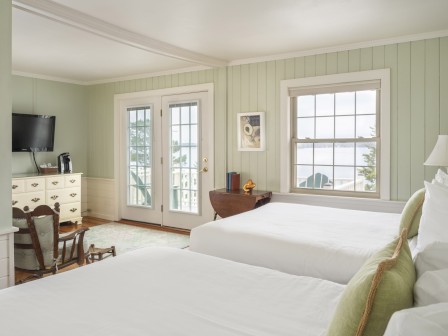 The image shows a cozy bedroom with two beds, a dresser, a TV, a spacious window, and French doors. The decor is light and airy.