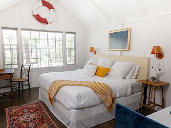 A cozy bedroom with a large bed, white walls, a window, a small desk with a chair, and nautical decor, including a lifebuoy on the wall.