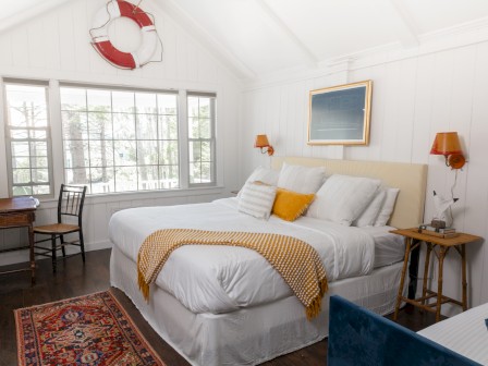 A cozy bedroom with a large bed, white walls, a window, a small desk with a chair, and nautical decor, including a lifebuoy on the wall.