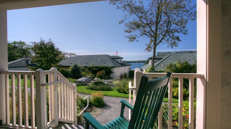 A green rocking chair on a porch overlooks a well-kept garden and buildings with a glimpse of the blue sky and sea in the background.