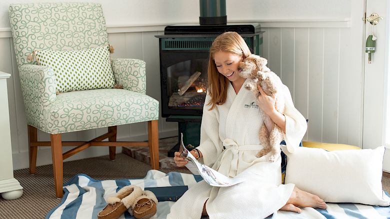 A woman in a white robe reads a magazine, sitting on a striped blanket with a small dog, near a fireplace and a cushioned chair with slippers beside it.