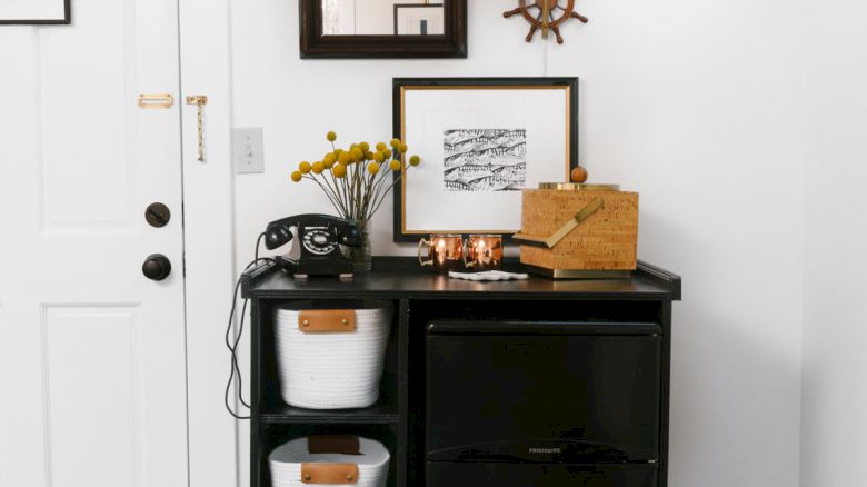 The image depicts a neat entryway setup with a small black shelving unit, three white storage bins, a rotary phone, a mirror, and decor on a blue rug.