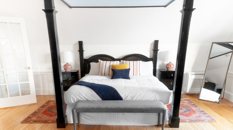 A neatly arranged bedroom with a four-poster bed, white bedding, a blue throw, a bench, two nightstands with lamps, and a large mirror.