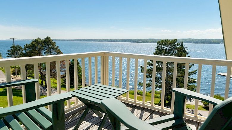 A balcony with green Adirondack chairs overlooks a serene body of water and trees under a clear blue sky.
