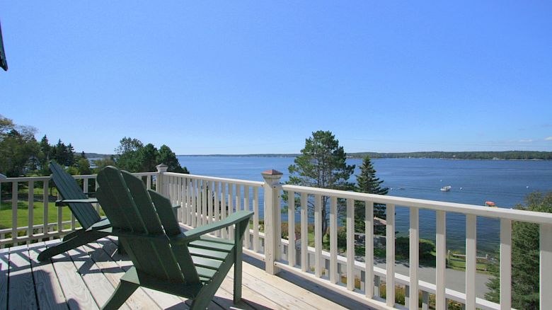 A serene waterside view from a wooden deck with two Adirondack chairs facing the water, surrounded by trees and a clear blue sky, ending the sentence.
