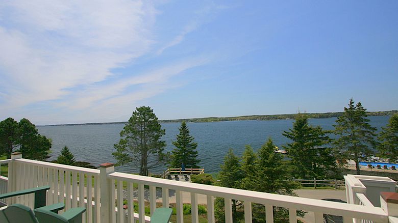 A scenic view from a balcony overlooks a serene lake with trees and a clear blue sky in the background. There are green chairs on the balcony.