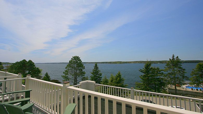 The image shows a scenic view from a balcony with chairs, overlooking a body of water and surrounded by trees under a partly cloudy sky.