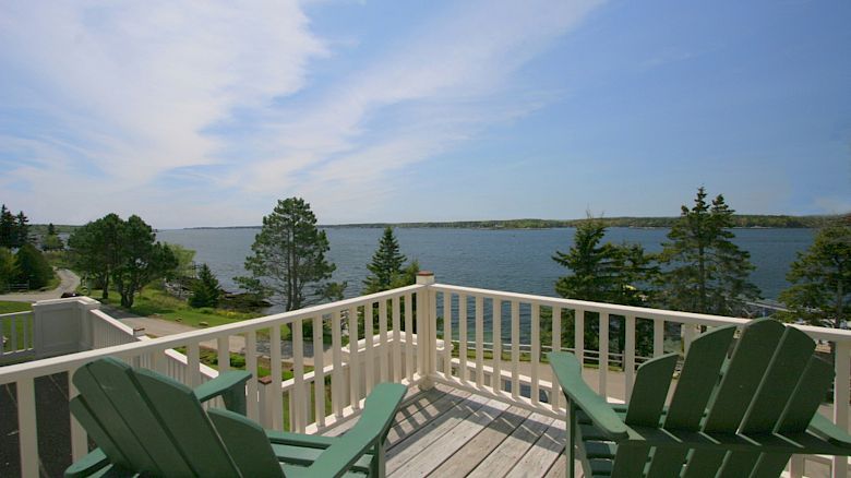 The image shows a balcony with two green Adirondack chairs overlooking a scenic lake with trees and a clear blue sky in the background.