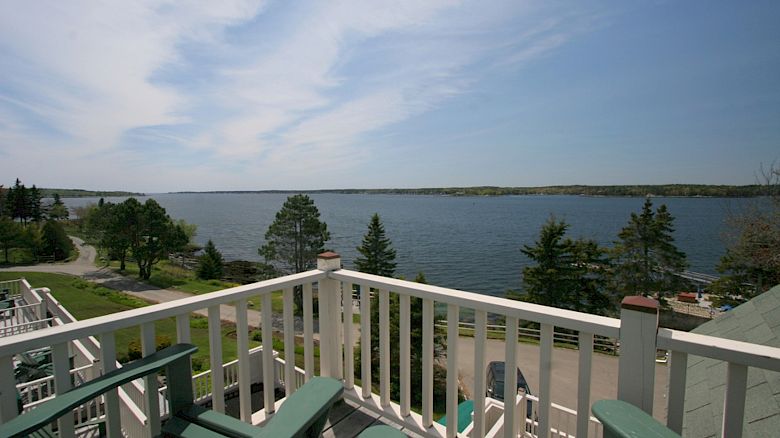 An outdoor balcony with green chairs overlooks a serene lake surrounded by trees, under a clear blue sky with wispy clouds.
