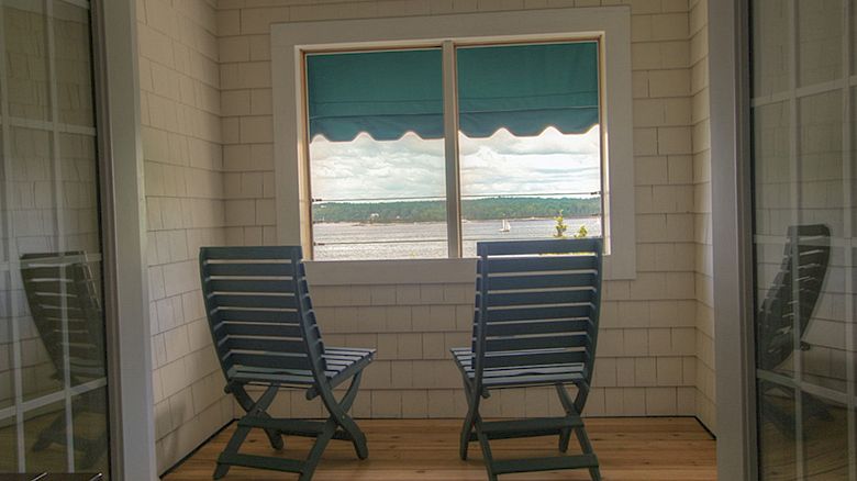 The image shows two wooden chairs facing a window with a view of a lake or river, with sailboats in the distance and a blue-green awning.