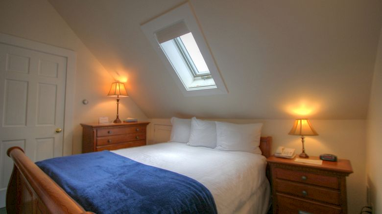 A cozy bedroom with a slanted ceiling, a skylight, a double bed with white linens and a blue throw, and lamps on wooden nightstands, ending the sentence.