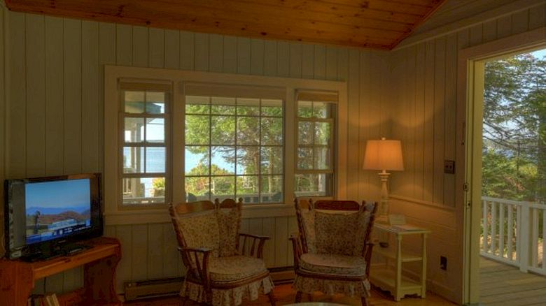 A cozy room with wooden interiors, two armchairs, a TV, and a lamp. A window and door offer views of trees and outdoor scenery, ending the sentence.
