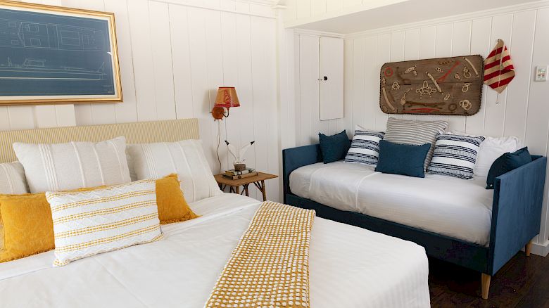 A cozy bedroom featuring a double bed and a daybed, decorated with white and yellow bedding, blue pillows, and nautical-themed wall art and accessories.