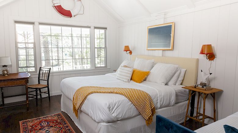 A cozy bedroom with a large bed, white walls, a window, and a desk. Decor includes a life preserver ring, a rug, and framed art.