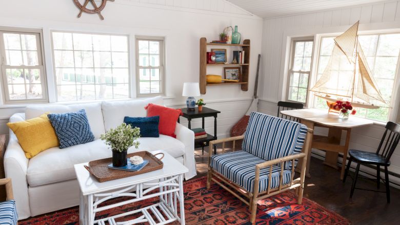 A cozy living room with nautical decor features a white sofa, striped chairs, a sailboat model, a red rug, and a bookshelf against white walls.