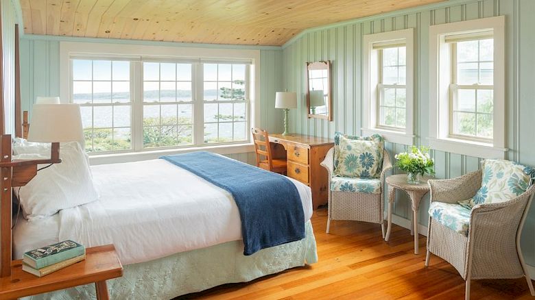 A cozy bedroom with wood flooring, light blue walls, large windows showing a lake view, a bed, seating area, desk, mirror, and lamps.