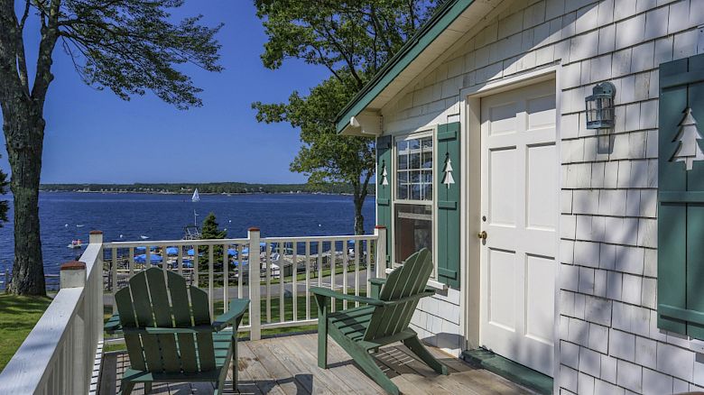 This image shows a porch with two Adirondack chairs facing a scenic lake or bay, adjacent to a house with green shutters and surrounded by trees.