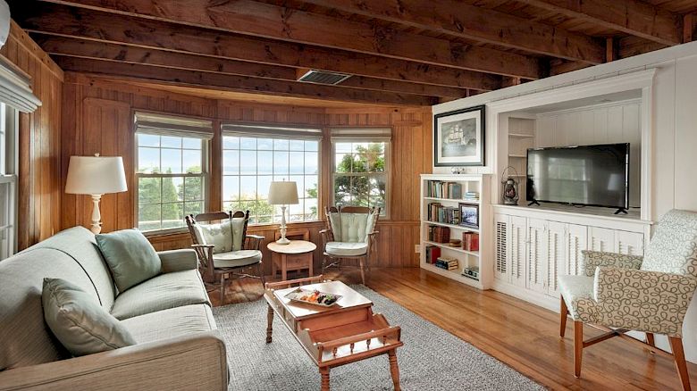 A cozy living room with wood-paneling, large windows, multiple chairs, a sofa, a TV, and a coffee table with a tray of snacks ends the sentence.