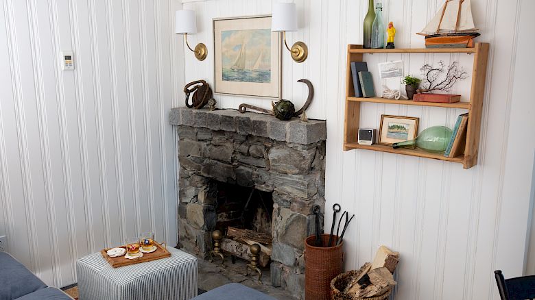 A cozy living room with a stone fireplace, nautical decor, a small ottoman with a tray, and a shelf displaying books, bottles, and small artworks.
