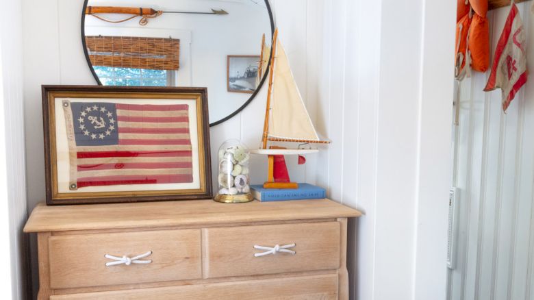 Wooden dresser with four drawers, a round mirror, framed American flag, and decorative sailboat. Wall-mounted lamp above. Room visible on the right.