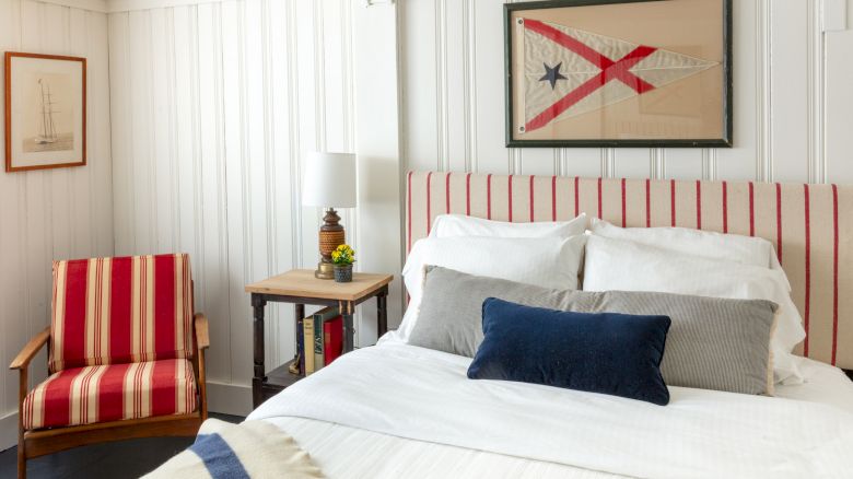 A cozy bedroom features a bed with striped red headboard and pillows, a striped chair, nautical decor, and framed art on white paneled walls.