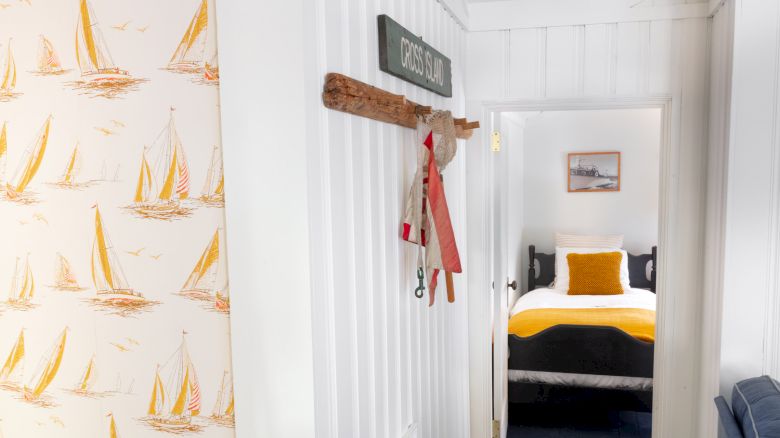 This image depicts a hallway leading to a bedroom with yellow accents, a nautical-themed wallpaper, and a decorative sign with life preserver.