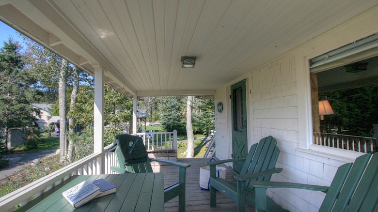 A cozy porch with green chairs and a table, an open book, and a view of the surrounding trees. A window and a door are on the right.
