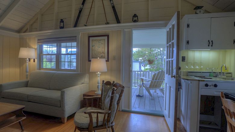 A cozy room has a sofa, rocking chair, lamps, and a kitchenette, opening to a veranda with Adirondack chairs.