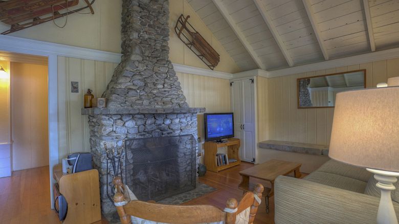 This image shows a cozy living room with a stone fireplace, wooden sleds on the wall, and a television. The room has high vaulted ceilings.