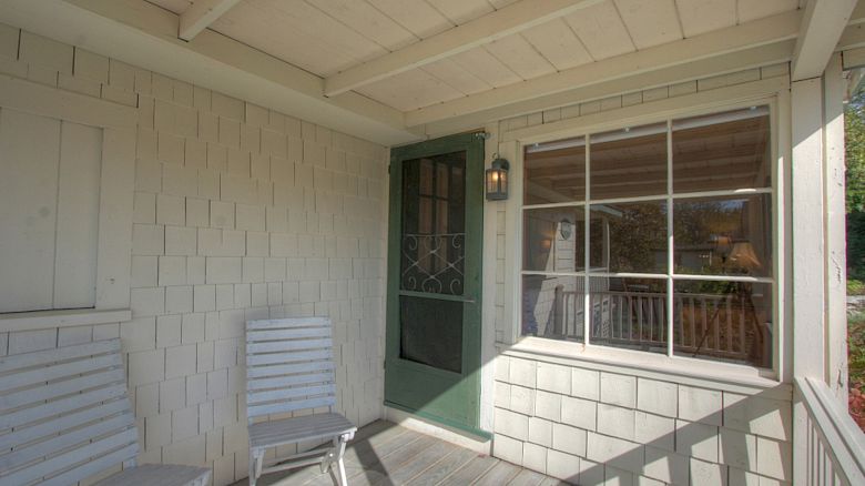 A porch with two white wooden chairs, a green door, and a large window with white framing. The area is shaded and has wooden flooring.
