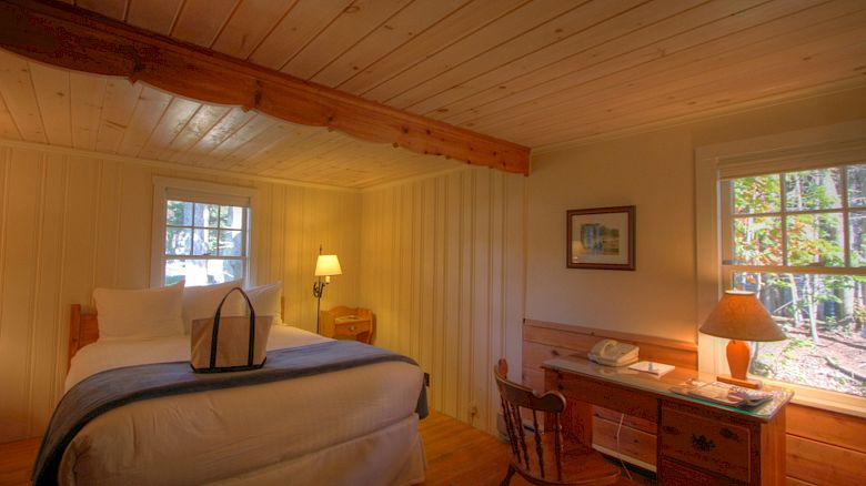 A cozy bedroom with wooden walls and ceiling, a bed with white linens, a desk with a lamp, and two windows letting in natural light.
