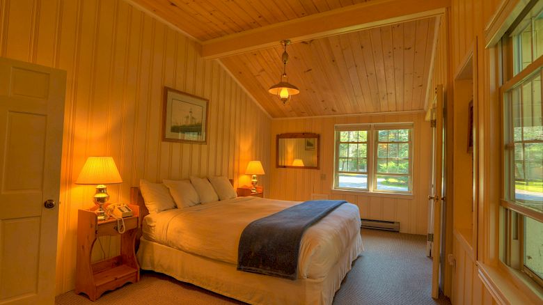 A cozy bedroom with wooden walls, a large bed, two nightstands with lamps, framed pictures, and windows letting in natural light.