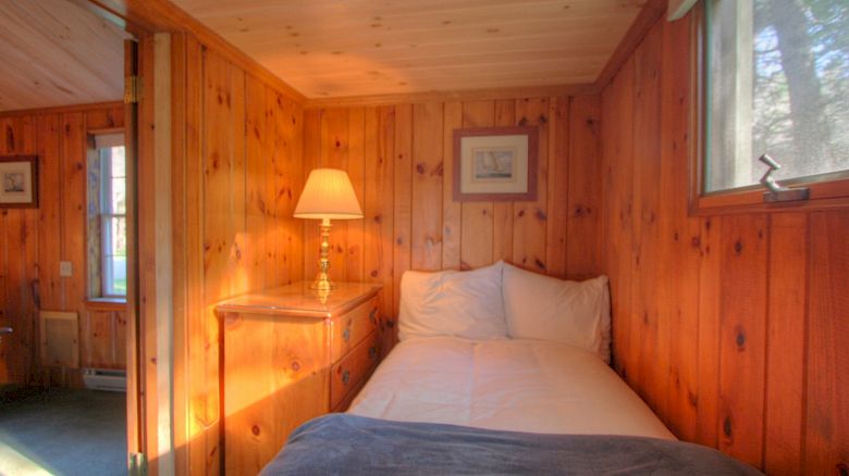 A cozy wooden room with a bed, lamp on a dresser, and picture frames on the wall. There is also a window letting in natural light.