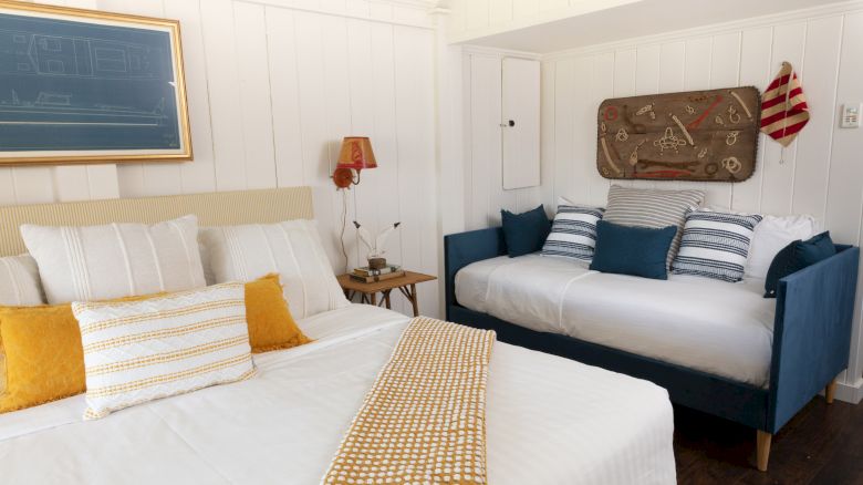 A cozy bedroom with a bed, a daybed, pillows in white and blue, yellow blankets, and a nautical-themed decor mounted on the wall.