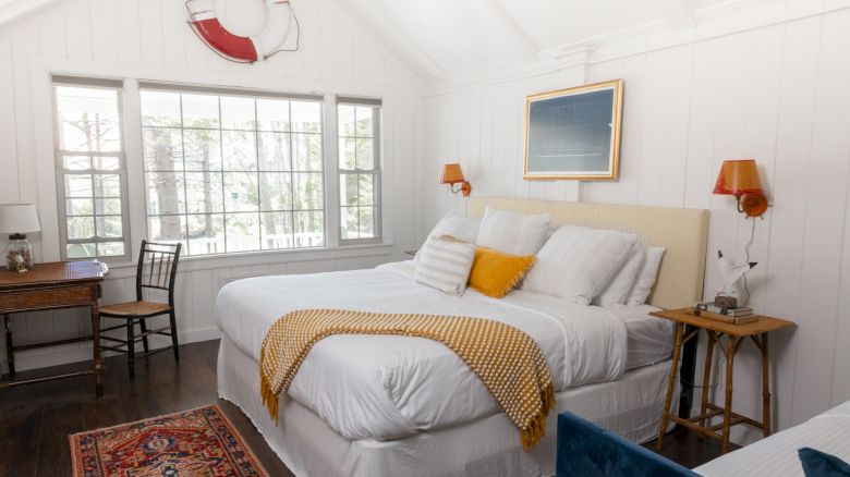 A cozy bedroom with a large bed, yellow pillow, and knitted throw. A window, life preserver decor, desk, chair, and side tables complete the look.