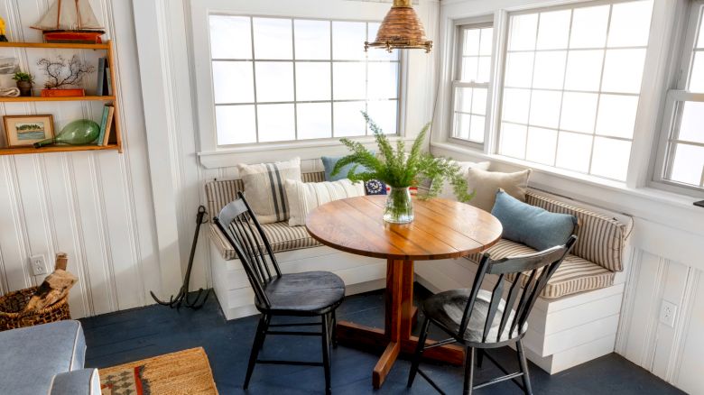 A cozy dining nook featuring a round wooden table, cushioned bench seating, two chairs, and a potted fern centerpiece, with plenty of natural light.
