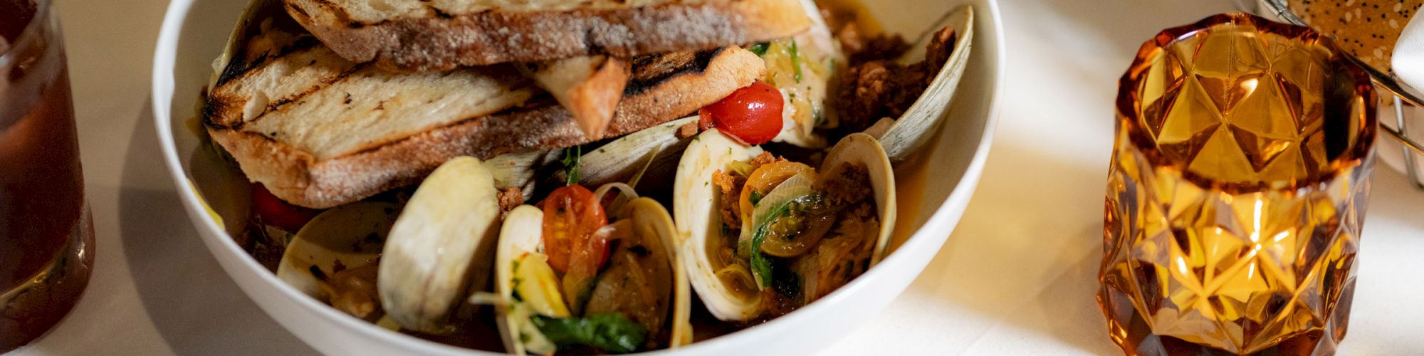 A bowl of seafood pasta with grilled bread, surrounded by drinks, a bread basket, and additional plates on a dining table.