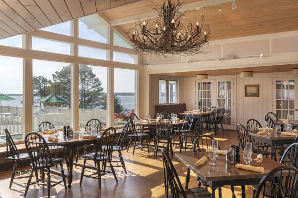 A well-lit, spacious dining room with large windows, scenic views, wooden tables, and chairs, featuring a rustic chandelier and set for guests.