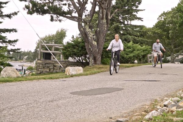 Two people are riding bicycles along a paved road with trees, grass, rocks around, and a waterfront in the background.