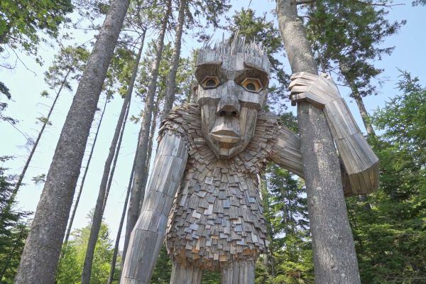 A large wooden sculpture of a humanoid figure stands among tall trees, holding onto one of them.