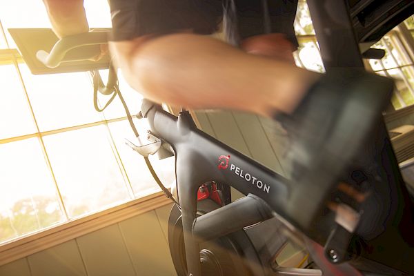 A person is riding a Peloton exercise bike indoors next to a window with sunlight streaming through, creating a bright and energetic atmosphere.