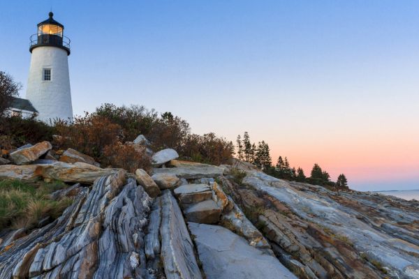 A lighthouse stands atop rocky cliffs with trees in the background and a clear sky at sunset.