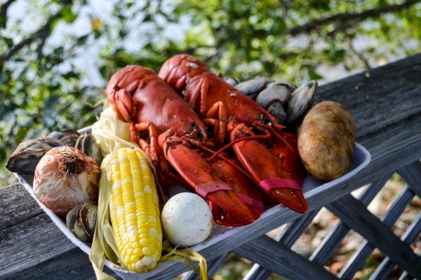 The image shows a platter with two lobsters, corn on the cob, clams, a potato, and possibly an onion or garlic bulb, all placed on a railing outdoors.