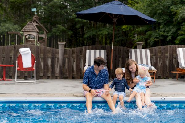 A family is sitting by the edge of a swimming pool, dipping their feet in the water, with lounge chairs and an umbrella in the background.